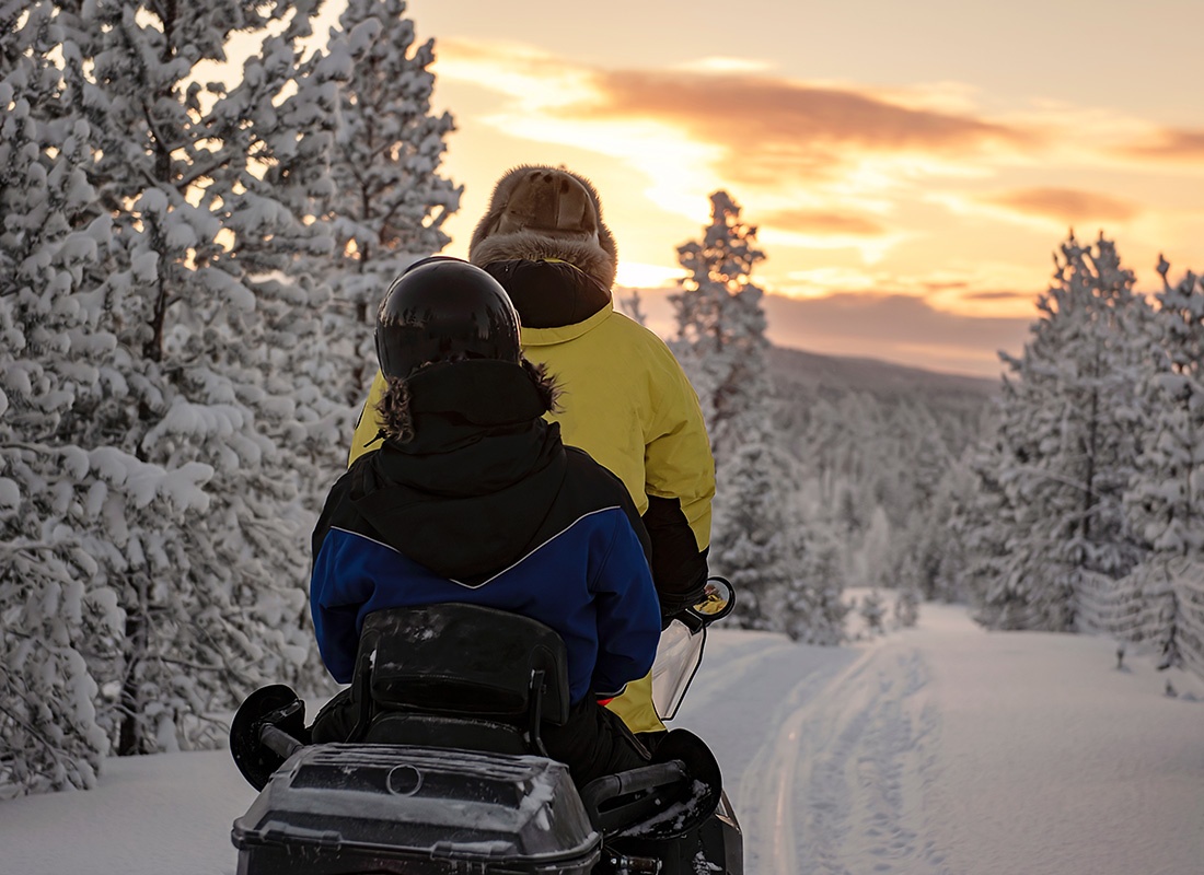 We Are Independent - View of Two People Riding a Snowmobile on a Snowy Path in the Forest Surrounded by Trees with a Colorful Winter Sunset Sky