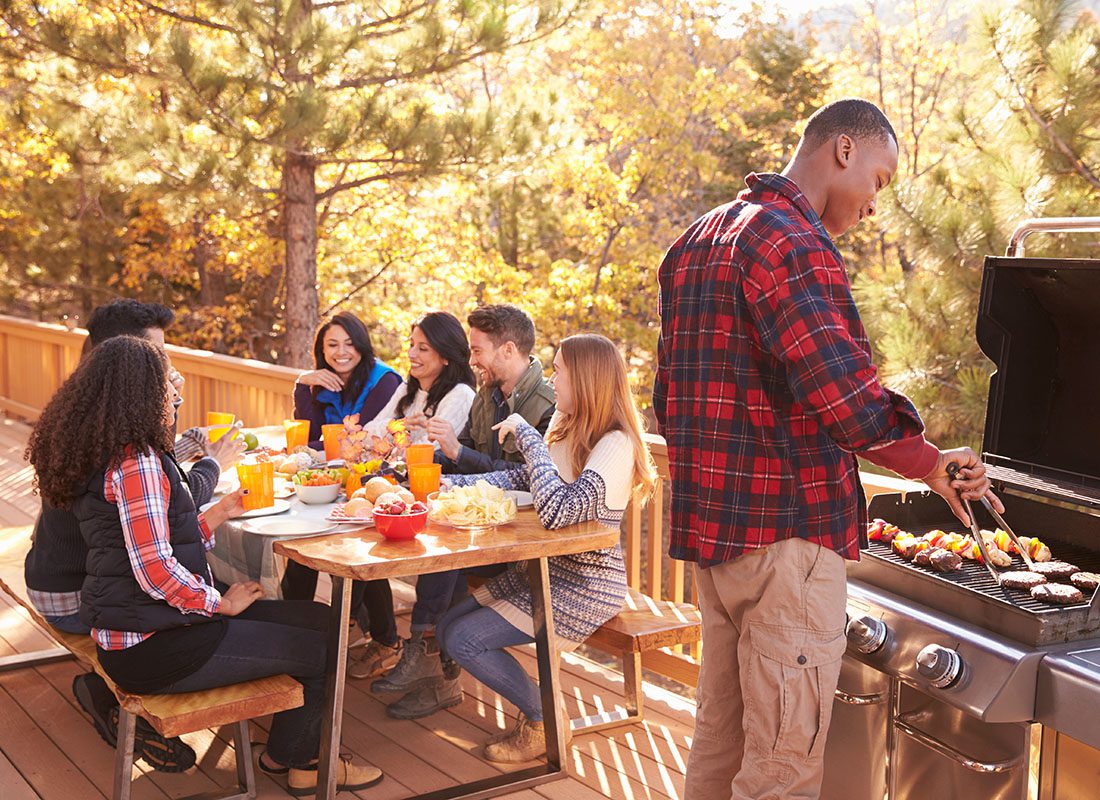 About Our Agency - Young Man Making Barbeque for his Group of Friends Sitting on a Wooden Table Outside on the Deck While on a Vacation Retreat in the Woods