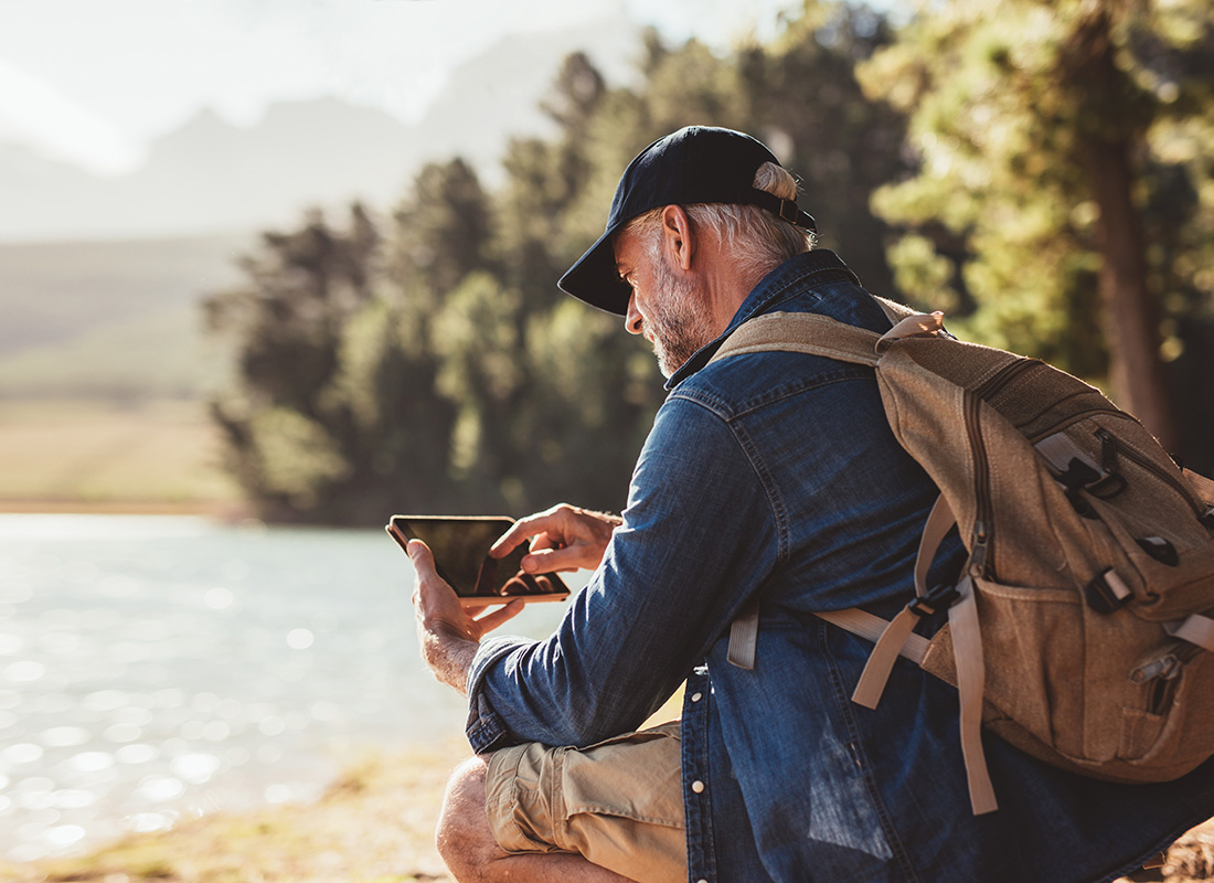 Read Our Reviews - Side View of a Mature Man Sitting by the Lake During a Hiking Trip While Using a Tablet on a Sunny Day